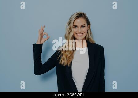 Fröhliche blonde Schülerin in legerer Kleidung, die ihre Zustimmung demonstriert und gleichzeitig eine gute Geste zeigt Stockfoto