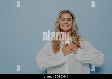 Glückliche dankbare Frau mit blondem welligen Haar in weißem Oversize-Shirt gestikulieren Wertschätzung und Liebe Stockfoto