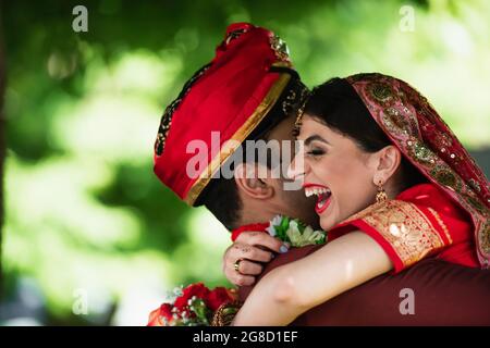 Indischer Mann im Turban umarmt glückliche Braut in traditionellem Kopftuch Stockfoto