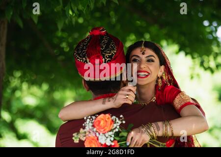 Rückansicht des indischen Mannes im Turban umarmt fröhliche Braut in traditionellem Kopftuch mit Blumen Stockfoto