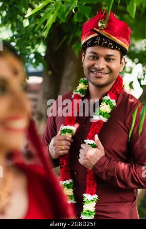 Glücklicher indischer Mann in Turban und Blumengirlande, der die verschwommene Braut ansieht Stockfoto