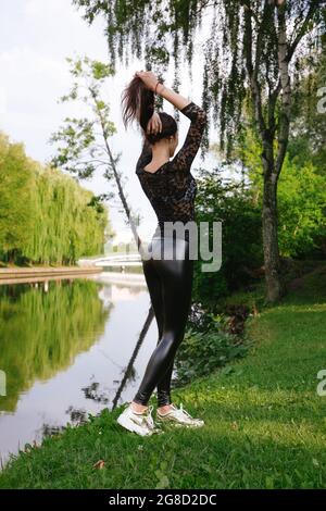 Hübsches Mädchen steht im grünen Park in der Nähe des Wassers; sie trägt schwarze Kleidung (transparentes Oberteil & glänzende enge Leggings) & goldene Turnschuhe. Sie zieht ihre Haare hoch Stockfoto