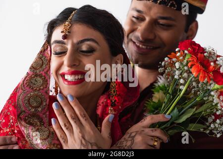 Verschwommener indischer Mann im Turban, der lächelnde Braut mit Mehndi, die Blumen auf Weiß hält, ansieht Stockfoto