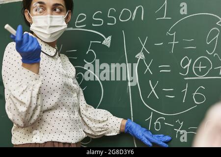 Männlicher Lehrer, der eine Gesichtsmask mit Latexhandschuhen trägt und der Klasse eine mathematische Formel erklärt Stockfoto