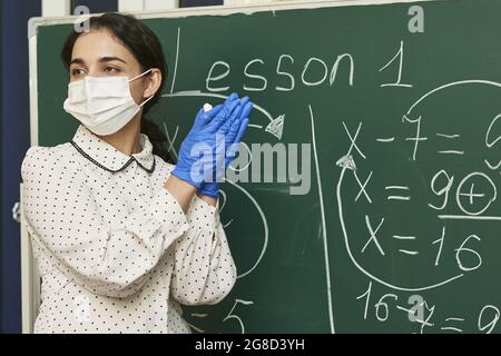 Die Lehrerin trägt eine Gesichtsmask mit Latexhandschuhen und erklärt der Klasse eine mathematische Formel Stockfoto