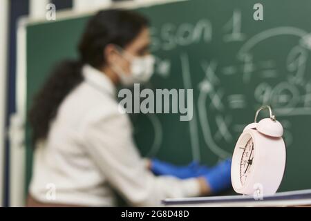 Wecker auf dem Tisch mit einer Lehrerin, die eine Facemask trug, während sie im Hintergrund unterrichtete Stockfoto