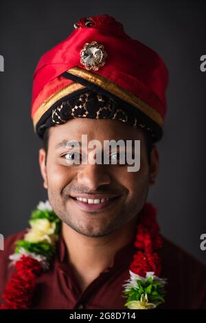 Positiver indischer Mann in Turban und Blumengirlande, der isoliert auf Grau posiert Stockfoto
