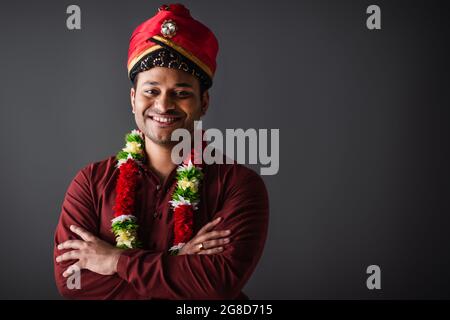 Lächelnder indischer Mann in Turban und Blumengirlande, der mit gekreuzten Armen auf grau isoliert steht Stockfoto