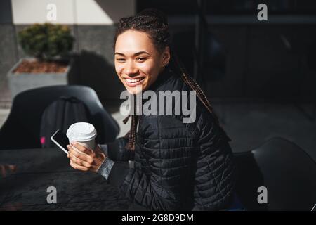 Eine fröhliche junge Frau, die mit einer Tasse Kaffee und einem Smartphone am Tisch sitzt Stockfoto