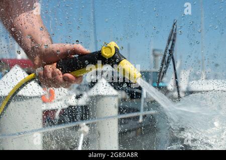 Hand halten Wasser-Spritzpistole. Mann, der Boot mit Druckwassersystem wascht. Blick vom Boot aus. Konzentrieren Sie sich auf Wassertropfen. Yacht-Wartungskonzept. Stockfoto