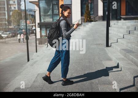 Fröhliche hübsche Touristin mit Kornreihen, die ihr Smartphone im Freien benutzt Stockfoto