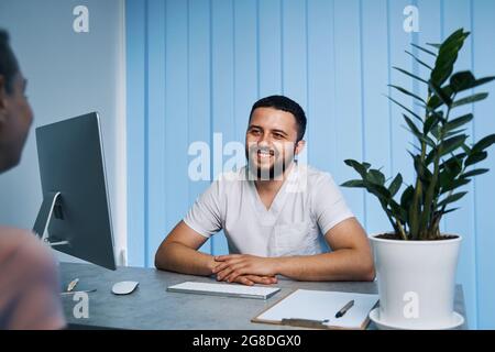 Glücklicher männlicher Arzt konsultiert weibliche Patienten und präsentiert das Ergebnis der Behandlung, während er am Arbeitsplatz sitzt Stockfoto