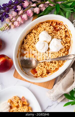 Frisch hausgemachter Nektarinkrümel mit Vanilleeis in Keramikform auf weißem Marmorboden. Draufsicht. Stockfoto