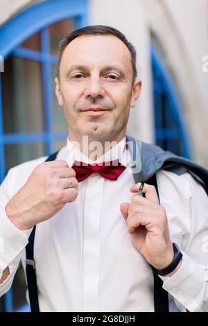 Nahaufnahme eines hübschen Mannes im Alter von 50 Jahren in formeller Kleidung, der seine rote Fliege anpasst und vor der Kamera lächelt. Verschwommener Blick auf das Stadtgebäude mit blauen Fenstern im Hintergrund Stockfoto