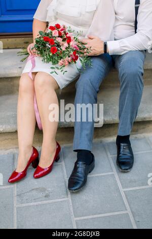 Nahaufnahme der Beine eines reifen Paares, eines Mannes und einer Frau in elegantem Anzug und Kleid, die auf einer alten Steintreppe mit Blumenstrauß im Freien auf der Straße der Stadt sitzen Stockfoto