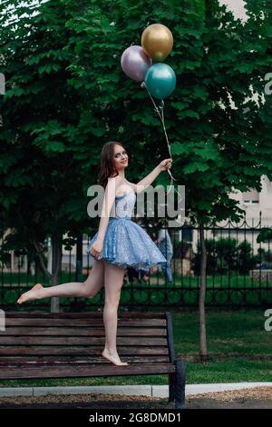 Eine junge Frau in einem blauen Kleid geht barfuß auf einer Holzbank und hält fliegende Ballons, so als ob sie fliegen würde Stockfoto