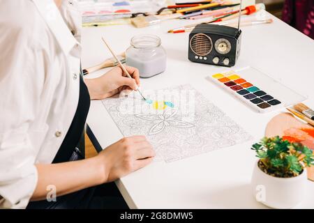 Junge Frau Malseite Antistress am Tisch drinnen, geistiges Wohlbefinden und Kunsttherapie. Frau malt eine Skizze, meditativen Prozess der Färbung von Seiten. Stockfoto