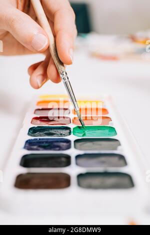 Nahaufnahme der Hand, die einen Pinsel hält und die Farben der Farben auf der Palette mischt. Konzept der Kunsterziehung, Kunstklasse oder Studio und Kunsttherapie. Farbe Stockfoto