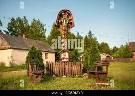 Schrein am Straßenrand und hölzernes, christliches Kreuz mit einigen Bänken und Zaun davor. Alte, kleine Dorfhäuser und landwirtschaftliche Gebäude Stockfoto