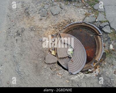 Zerbrochene Metallschachtabdeckung aus Eisen auf leerer Luke führt zum städtischen Abwassersystem. Draufsicht Stockfoto