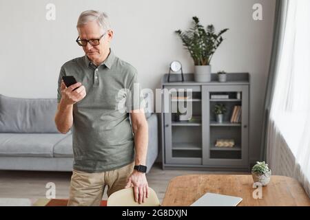 Minimales Porträt eines modernen älteren Mannes, der sein Smartphone benutzt, während er im Inneren des Hauses steht und Platz kopiert Stockfoto