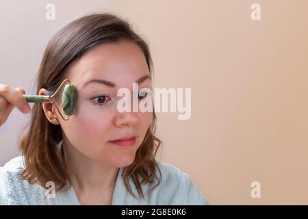 Hübsche Frau hält Jade Gesichtswalze und macht Gesichtsmassagen. Hauspflegekonzept für zu Hause Stockfoto