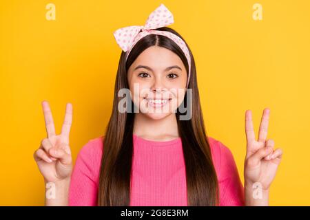 Foto von entzückenden niedlichen Schulmädchen tragen rosa T-Shirt zeigt zwei V-Zeichen lächelnd isoliert gelbe Farbe Hintergrund Stockfoto