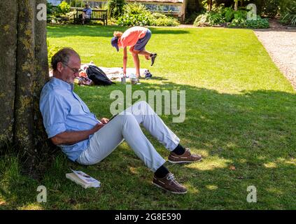 Ein Mann liest, der sich im Chelsea Physic Garden an einem großen Baum lehnt, während eine Frau im Hintergrund versucht, sich zu balancieren, um ihre Schuhe anzuziehen. Stockfoto