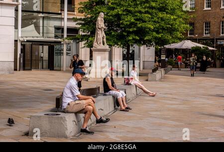 Die Menschen sitzen in Sommerkleidung an der Statue von Sir Hans Sloane auf dem Sloane Square im Zentrum Londons. Sloane ist mit Sklaverei verbunden. Stockfoto