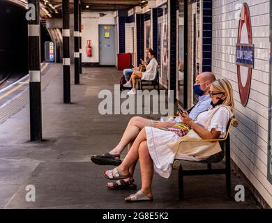 Ein Paar, das auf einer Bank auf dem Bahnsteig sitzt und in Masken auf einen U-Bahn-Zug im Zentrum Londons wartet Stockfoto