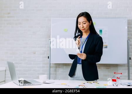 Zufriedene asiatische Frau in formellem Outfit mit Abzeichen lächelnd und Papier zu lesen, während sie am Tisch mit Marketingplan und Laptop im Büro steht Stockfoto