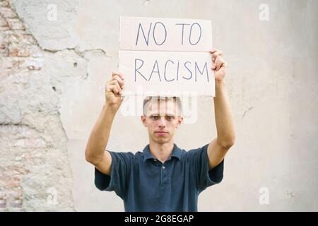 Der kaukasische Typ kam mit einem Plakat in den Händen heraus, um gegen Rassismus zu protestieren. Der Auftritt des jungen Mannes steht vor dem Hintergrund einer Zementwand mit Pappe in der Hand, auf der es Nein zum Rassismus heißt Stockfoto