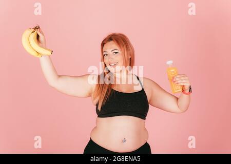 Eine diätetigeweise und entgiftende, lächelnde, dicke Frau im Sportoutfit hält auf rosa Studiohintergrund einen Haufen Bananen und eine Flasche Saft. Stockfoto