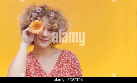 Junge attraktive Mädchen posiert mit orange Gerbera Gänseblümchen Blume. Bedeckt ihr Auge mit Gänseblümchen. Isoliertes Mädchen mit hellgelben Hintergrund Studio. Glückliches Mädchen lächelt und trägt Blumen in ihrem lockigen Haar. Stockfoto