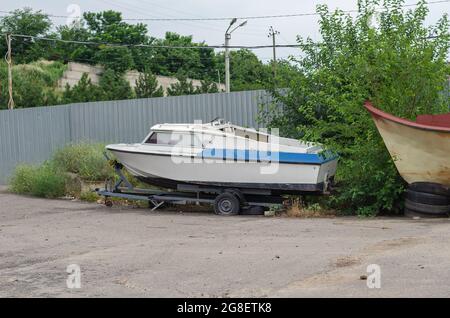 Ein altes baufälliges Fischerboot auf einem Anhänger vor einem grauen Zaun. PKW-Anhänger mit einem Platten Reifen auf dem Parkplatz. Tagsüber. Draußen. Stockfoto