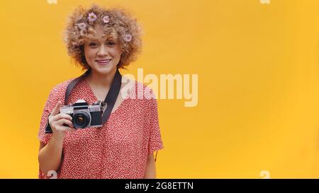 Mädchen hält eine Vintage-Kamera in einer fröhlichen Stimmung. Eine Kamera, die um ihren Hals hängt. Hellgelber Hintergrund. In einem lässigen rosafarbenen Hemd mit Blumen im Haar gekleidet. Fotokonzept. Stockfoto