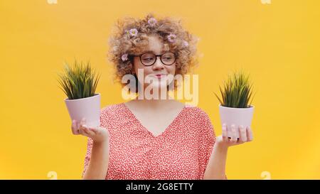 Fröhliches Mädchen, das zwei kleine Topfpflanzen in den Händen hält. Mädchen ist in einem lässigen rosa Top gekleidet und trägt nerdy Brille. Isoliert über hellgelben Hintergrund Studio. Stockfoto