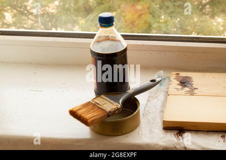 Set zum Bemalen von frisch gehobeltem Holz auf dem Fenster. Eine Flasche brauner Holzfleck und ein Pinsel auf dem Farbbehälter. Grüne Bäume an sonnigen Tagen Stockfoto