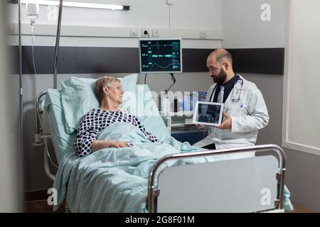 Der Arzt sitzt neben einem älteren Mann und erklärt die Diagnose eines Körpertraumas und zeigt Röntgenaufnahmen auf einem Tablet-pc im Krankenhauszimmer. Reifer Patient in der Klinik atmet es Hilfe aus Sauerstoffmaske. Stockfoto