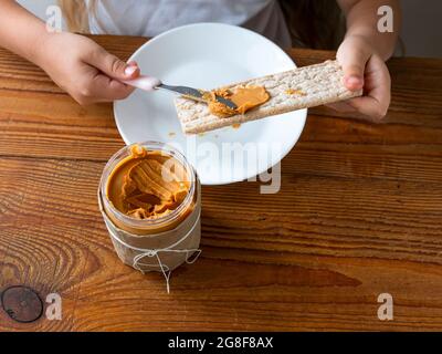 Kind Verbreitung Erdnussbutter auf Knäckebrot Essen Sandwich Holztisch Haus kitchen.School Mädchen mit Brot Scheibe Vollkorn Snack.Kid Kochen Frühstück Stockfoto