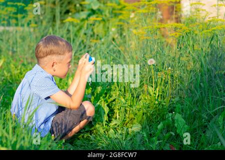 Ein süßer Vorschuljunge mit einer hübschen Frisur in einem blauen Hemd fotografiert an einem heißen Sommertag grüne Pflanzen. Selektiver Fokus. Hochformat Stockfoto