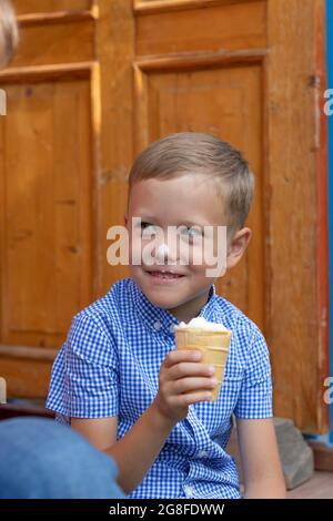 Zufriedener fröhlicher Junge in einem blauen Hemd isst an einem sonnigen Sommertag Eis auf der Veranda eines Hauses in einem Dorf. Selektiver Fokus. Hochformat Stockfoto