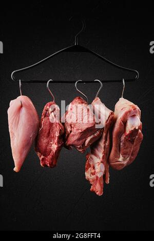 Verschiedene Schnitte von rohem Fleisch auf Haken auf Kleiderbügel über schwarzem Hintergrund Stockfoto