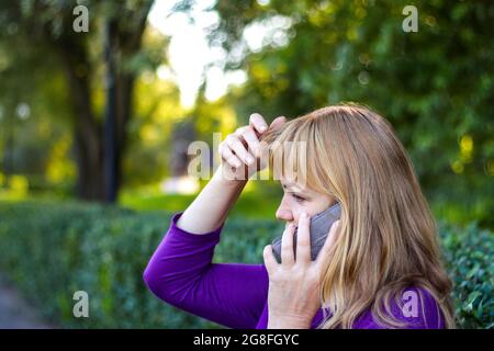 Unschärfe kaukasisch blonde Frau sprechen auf dem Telefon draußen, draußen. 40-jährige Frau in lila Bluse im Park berühren ihre Haare. Ernsthafte Person, die verwendet Stockfoto