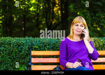 Unschärfe kaukasisch blonde Frau reden, sprechen am Telefon draußen, im Freien. 40-jährige Frau in lila Bluse im Park auf der Bank. Erwachsene, weiblich pro Stockfoto
