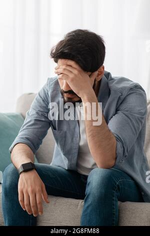 Konzept der männlichen Depression. Verzweifelter arabischer Mann, der seine Stirn berührt, Probleme hat, zu Hause auf dem Sofa sitzt, Ernte Stockfoto