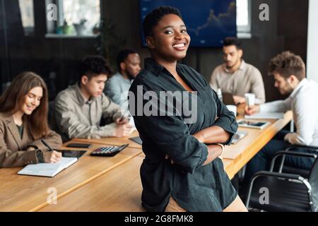 Lächelnde afroamerikanische Geschäftsfrau, die auf dem Schreibtisch sitzt und posiert Stockfoto