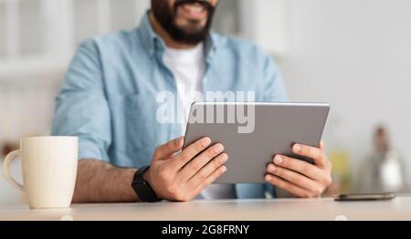 Arbeiten Sie von zu Hause aus. Nicht erkennbarer junger bärtiger Mann, der ein digitales Tablet hält, am Tisch in der Küche sitzt, Ernte Stockfoto
