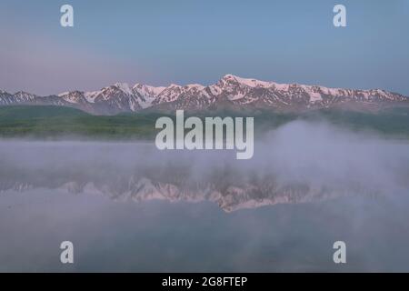 Erstaunlicher zarter Sonnenaufgang über den mit Schnee und grünem Wald bedeckten Bergen, Nebel über dem See und Reflexionen im Wasser im Sommer. Altai, Russi Stockfoto