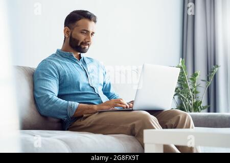 Ernsthafter junger Mann aus dem Osten, der am Laptop arbeitet, während er zu Hause im Hotel auf dem Sofa sitzt Stockfoto